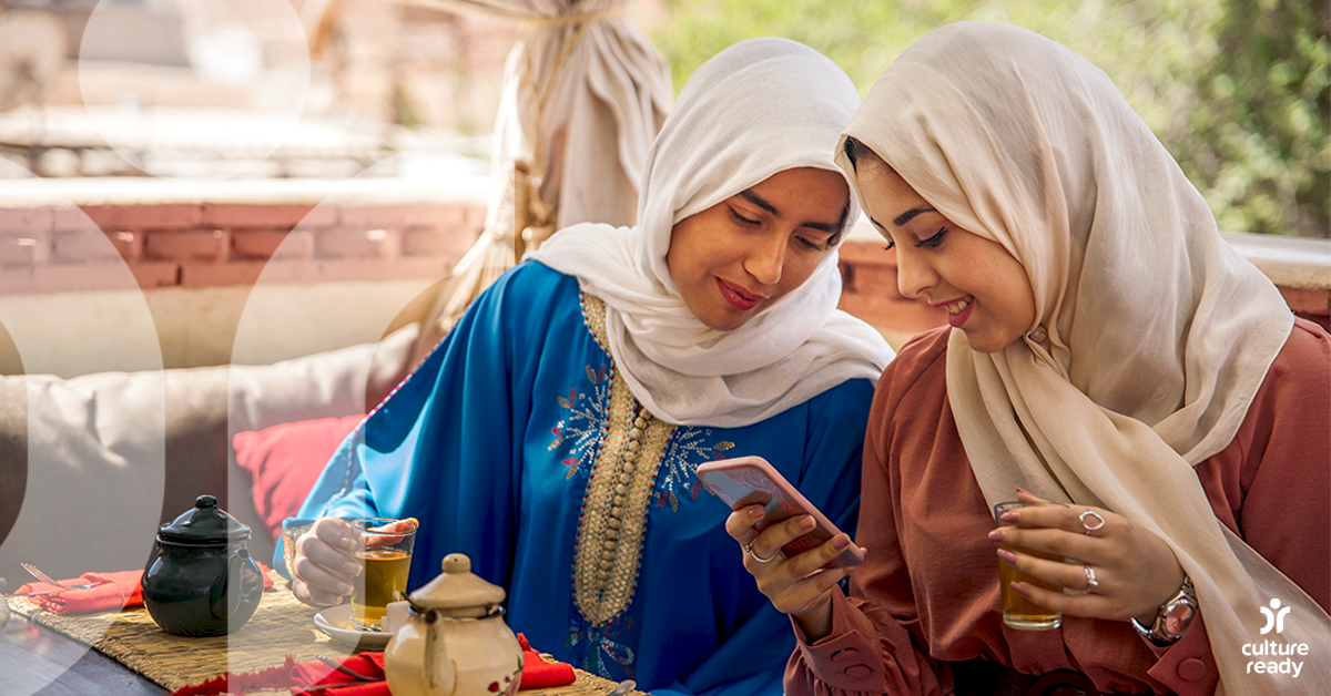 Two women in hijabs looking at a phone. One woman is holding a glass of tea.
